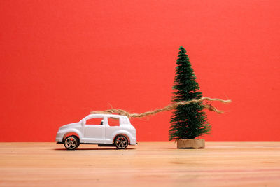 Close-up of small christmas tree tied with toy car on wooden table against red background