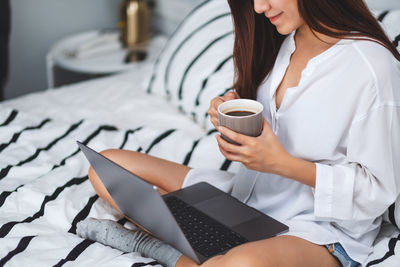 Midsection of woman holding coffee cup sitting on bed