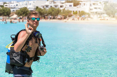 Portrait of shirtless young man wearing sunglasses showing ok sign while standing at beach