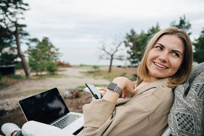 Smiling young woman with technologies looking away while sitting on chair during vacations