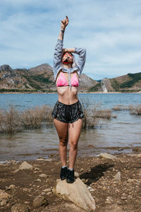 Young woman on the shore of a lake in spain in a bikini and shorts. standing in a relaxed position with her arms up