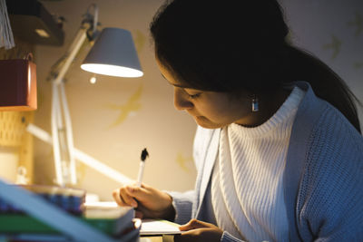 Girl writing in book while doing homework under illuminated desk lamp at home