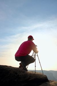 Photographerin red jacket and red baseball cap stay with camera on tripod on cliff works