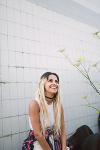 Smiling young woman looking away while sitting on tire against tiled wall