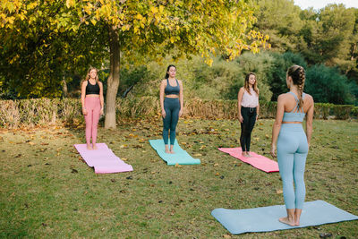 Group of young females standing in mountain pose while practicing yoga together with instructor in green park