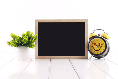 Close-up of blank blackboard with potted plant and alarm clock on table against white background