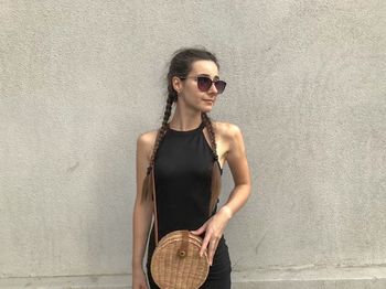 Portrait of young woman wearing sunglasses standing against wall