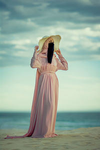 Front view of muslim woman wearing hat, niqab,  and hijab standing at the beach against cloudy sky