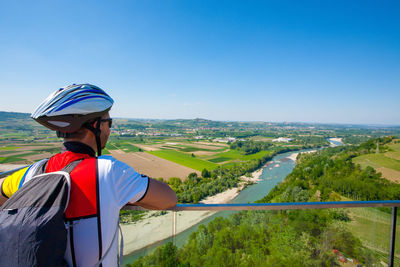 Rear view of man leaning on railing against landscape and blue sky