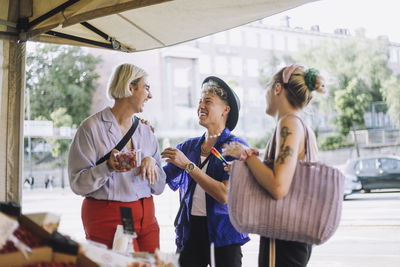 Happy young man talking with non-binary friend holding container of fruit