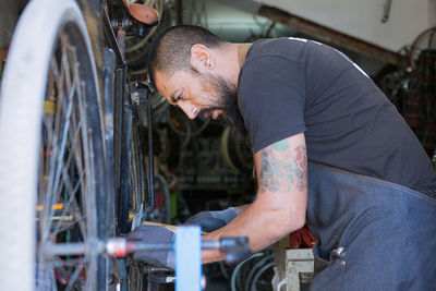 Concentrated male mechanic with beard and tattoos in gloves repairing bicycle in modern workshop