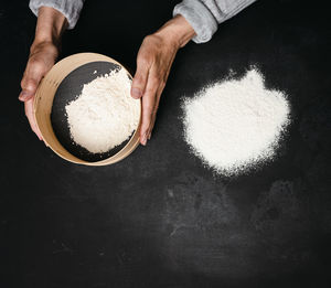 Two female hands hold a round wooden sieve with wheat flour on a black table, top view