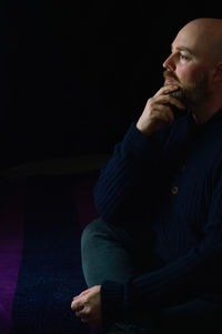 Young man looking away while sitting against black background