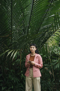 Young woman using mobile phone while standing by plant