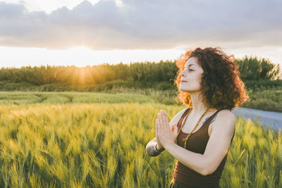 Woman meditating while standing at farm against cloudy sky during sunset