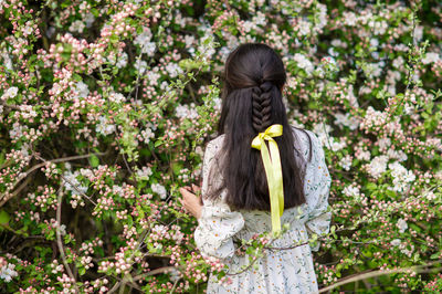 A young girl turned her back near a blooming cherry tree. yellow ribbon in a braid
