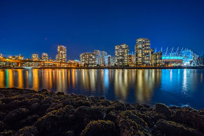 Illuminated buildings by river against blue sky at night