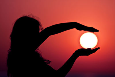 Optical illusion of silhouette woman holding sun against orange sky during sunset