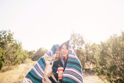 Portrait of couple with dog wrapped in blanket against trees against clear sky
