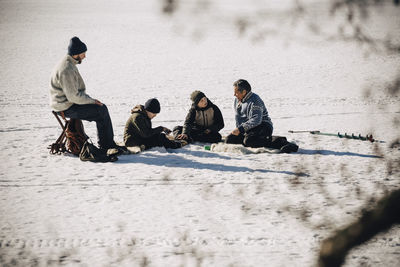 Mature men with sons sitting on snow while ice fishing during sunny day