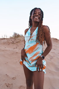 Low angle of happy african american female in summer dress standing on sandy beach against sunset sky and looking at camera