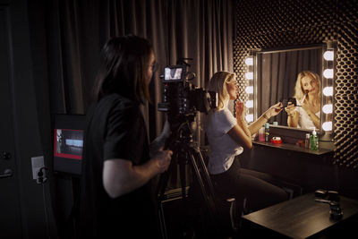 Man filming woman applying make-up in front of mirror