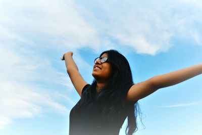 Low angle view of young woman with arms outstretched standing against sky