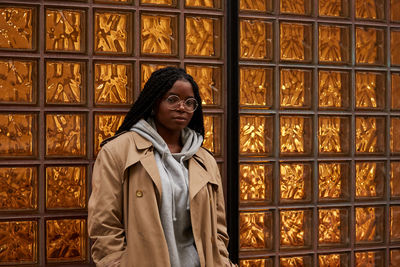 Pretty african american female with braids in casual outfit looking at camera while leaning on ornamental glass wall