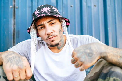 Young tattooed male hipster in informal clothing sitting on ground near metal fence and looking at camera