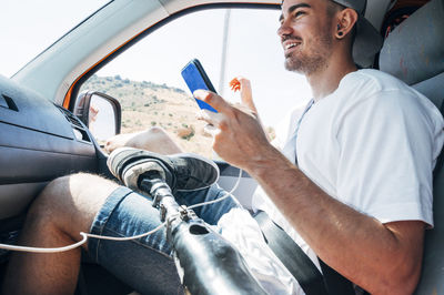 Smiling young man with leg prosthesis sitting in camper van using smartphone