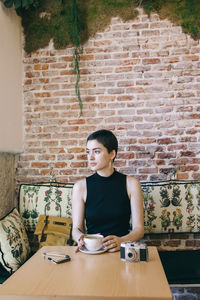 Young woman sitting by table against brick wall