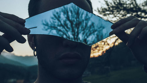 Close-up of man holding broken mirror with reflection of bare tree on it