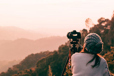 Rear view of mid adult woman photographing while standing on mountain against sky during sunset