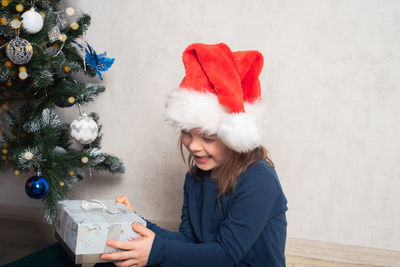 Happy girl in santa hat sits under the christmas tree and holds a gift. 