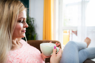 A blonde woman holds a cup of coffee while sitting in a chair.