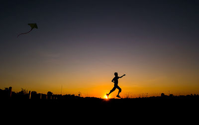 Silhouette boy flying kite while running on field during sunset