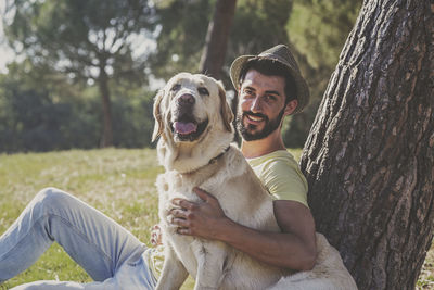 Portrait of mid adult man with dog relaxing on grassy field by tree trunk in park