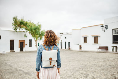 Back view of anonymous female with backpack walking on pavement against white houses and cloudy gray sky on town street in fuerteventura, spain