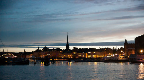 Illuminated buildings by river against sky at sunset