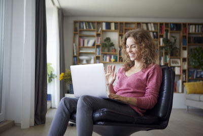 Smiling woman waving hand to video call on laptop while sitting at home