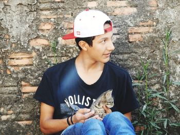 Teenage boy with cat looking away while sitting against wall