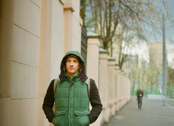 Portrait of young man in green jacket standing against building