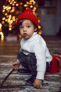 Child in a white sweater and a red knitted hat stands next to the christmas tree at home