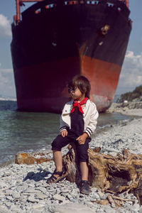 Fashionable boy child with long hair sits on a log next to a large ship that ran 