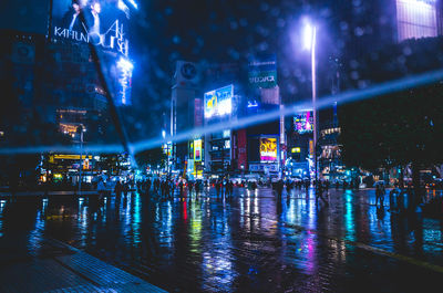 Illuminated buildings by wet road during rainy season at night