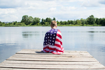 Rear view of young man holding american flag while sitting on pier against sky