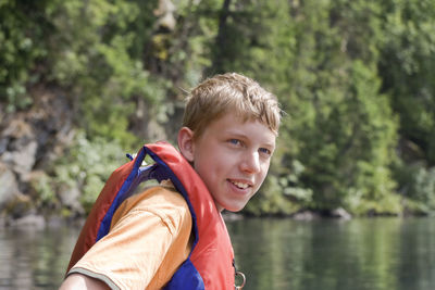 Teenage boy in life jacket looking away while sitting by lake