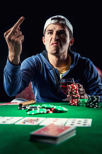 Young man showing obscene gesture while playing poker against black background