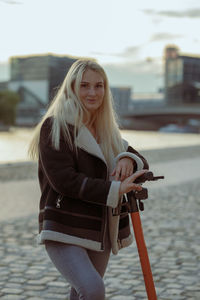 Portrait of young woman standing with push scooter on footpath in city