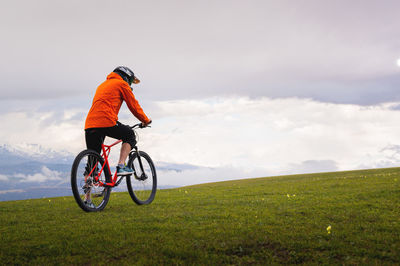 Man stands with a bicycle on a green lush meadow, against the backdrop of mountains and a cloudy sky